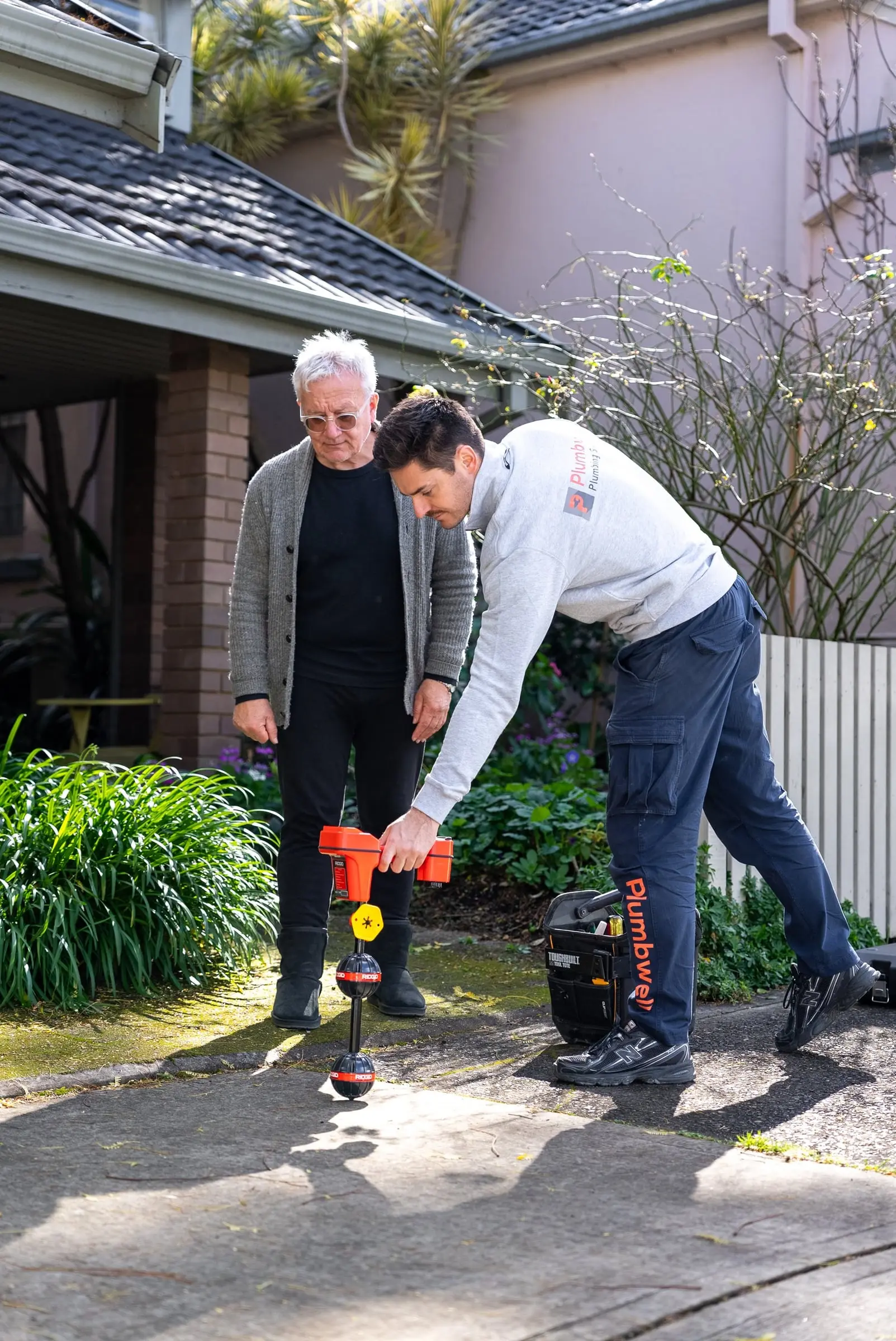 Plumber tracing an underground pipe with a locator while the owner watches