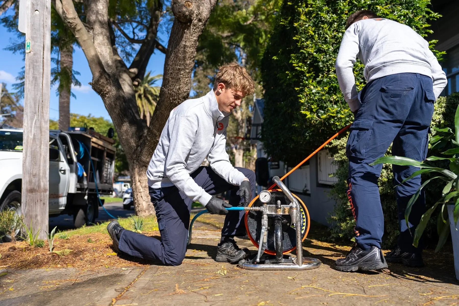 Two plumbers running a drain cleaning machine on the footpath beside their ute